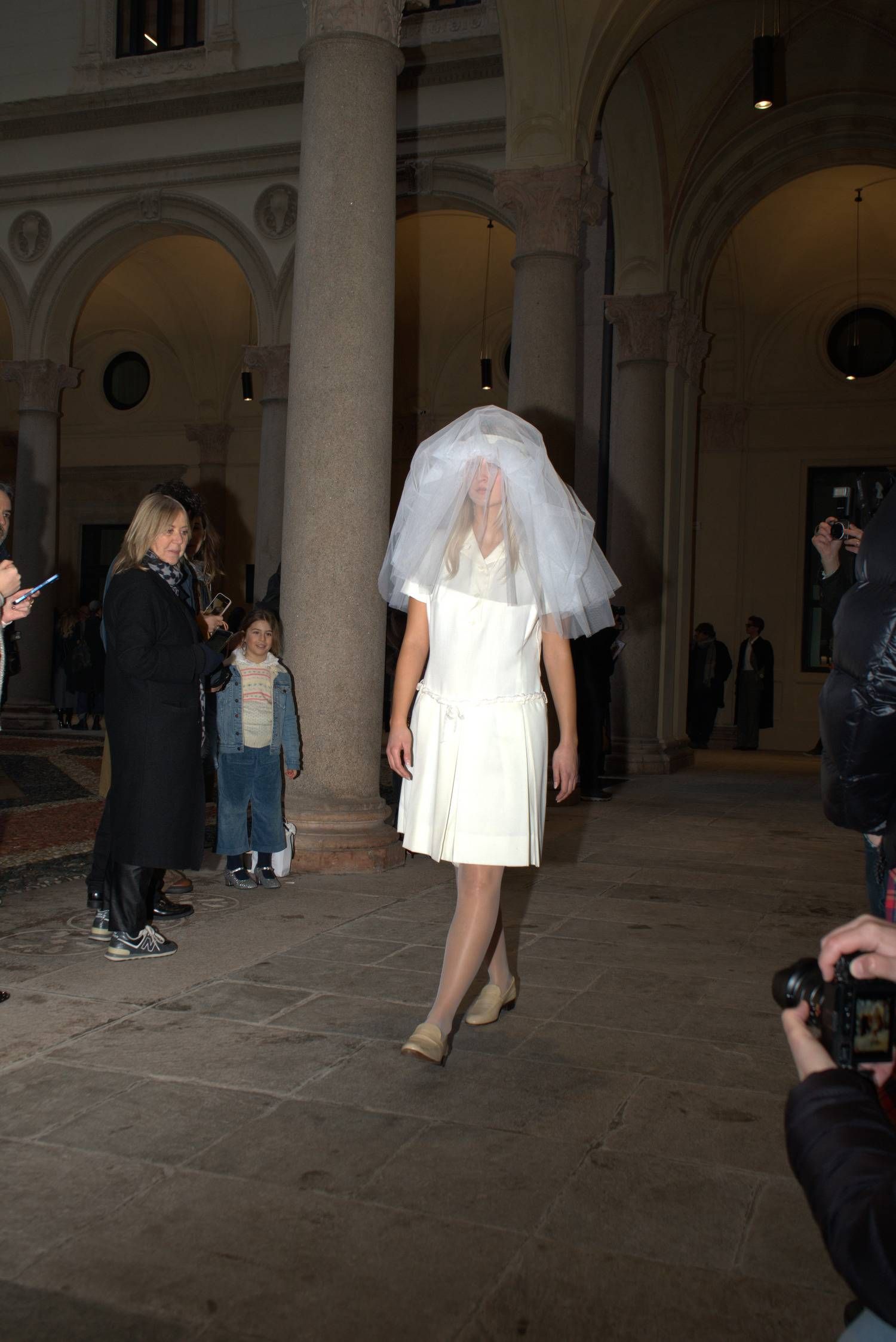 A suspended bridal figure: a simple white dress softened by a translucent veil, evoking rites of passage, domestic spirituality and memory reassembled through restraint.