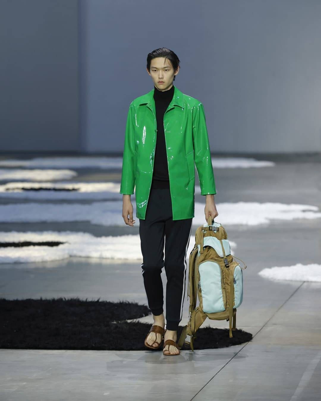 Prada menswear look featuring a glossy green coat worn over a black turtleneck, slim black trousers, leather sandals and a technical backpack